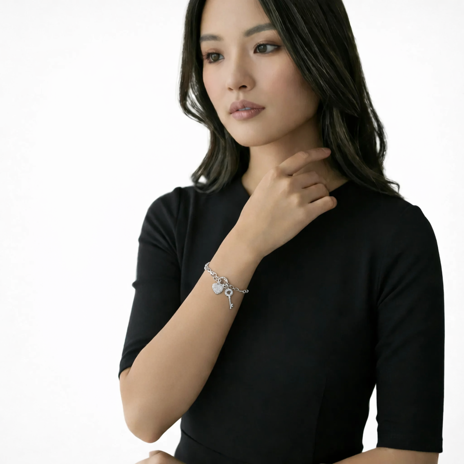 Model shot presents a polished charm bracelet with heart and key dangles resting against a black short-sleeve dress as the hand grazes the collarbone.